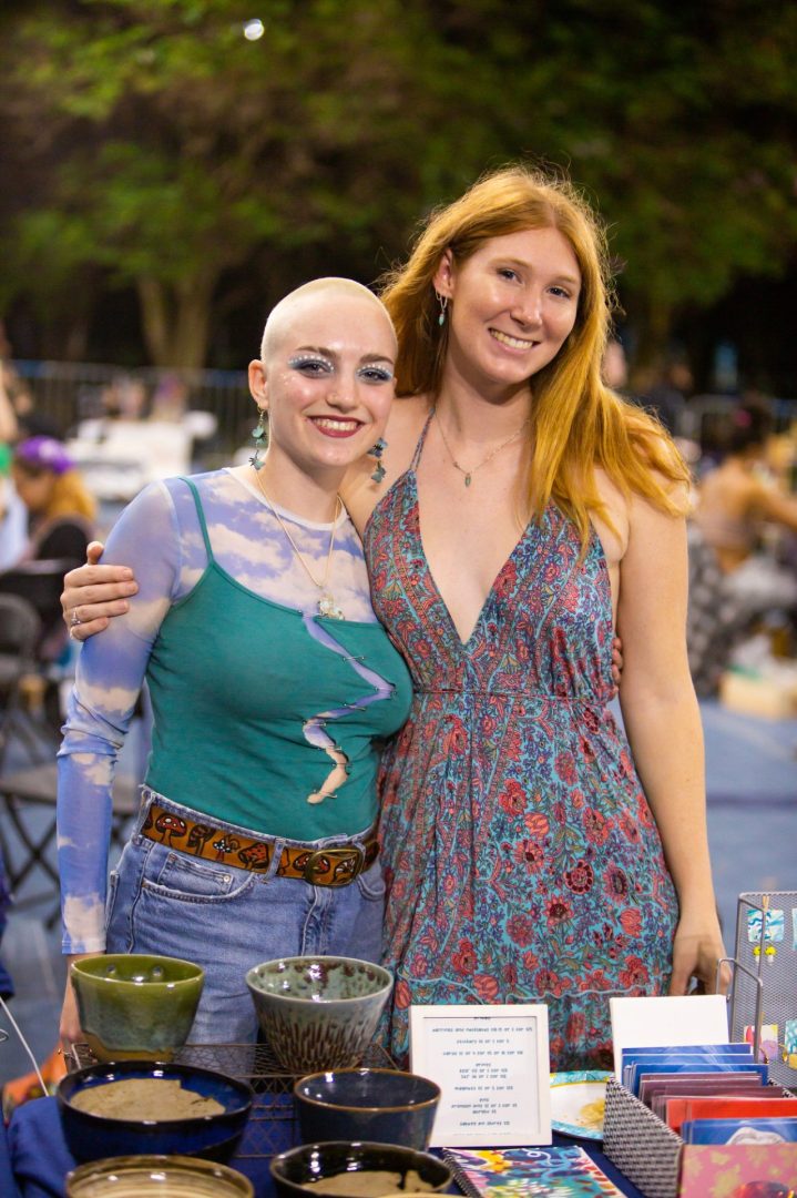 Two students shoulder to shoulder with pottery on table in front of them