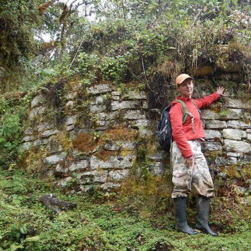 Professor in boots walking along old stone building in the woods