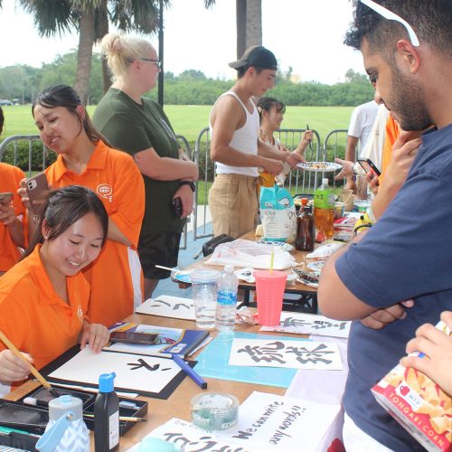 A student in an orange shirt paints words in Japanese while other students look on