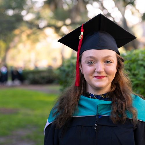 Student in graduate cap and gown in quad with other graduates walking by