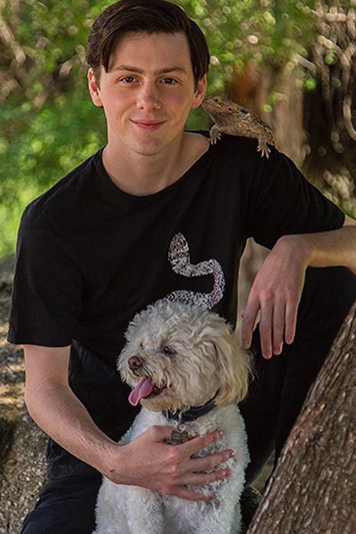 Student seated in a tree with a dog in lap and reptile on shoulder