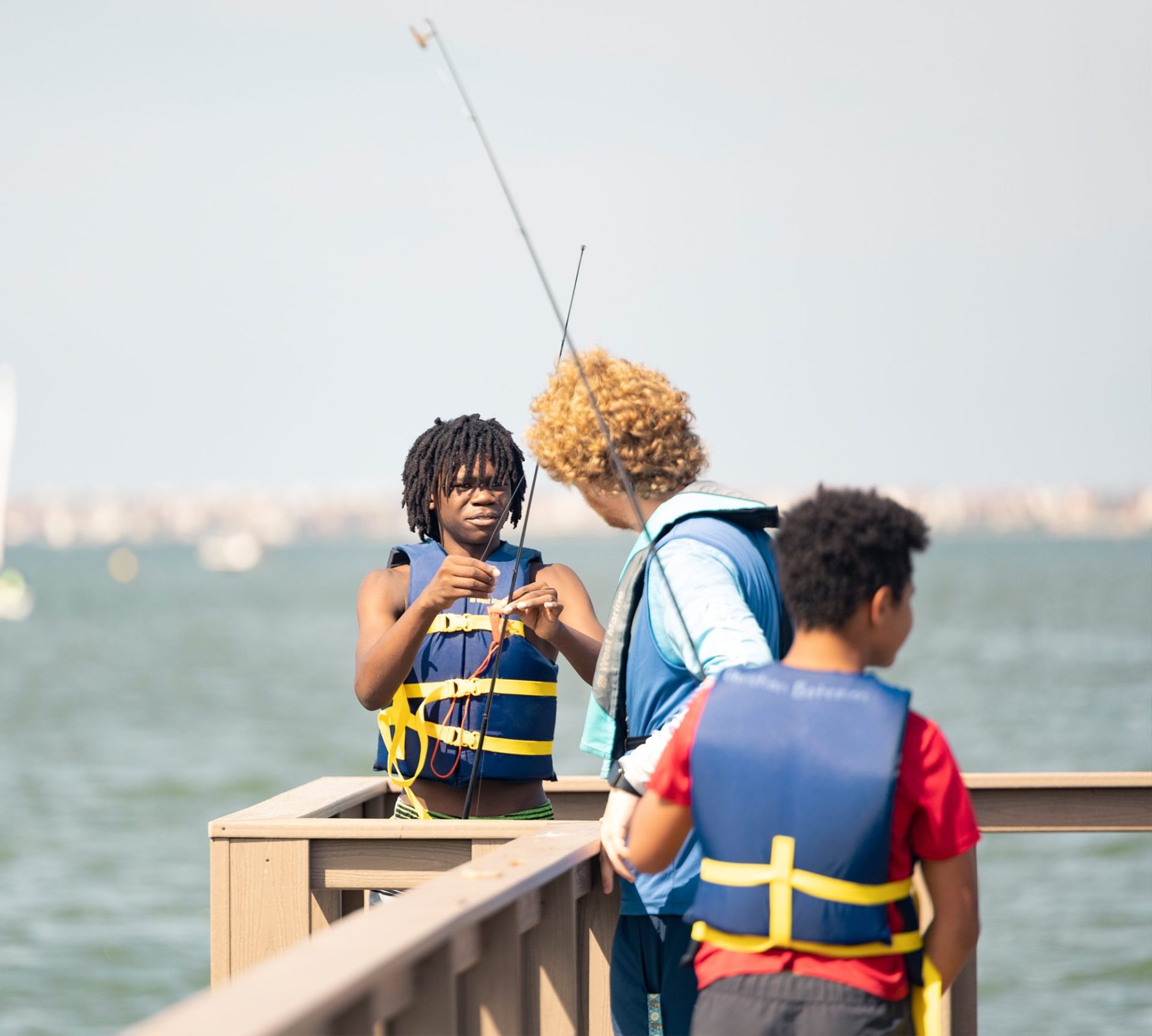 Two children wearing life vests cast fishing lines at the Eckerd College pier as an instructor observes.