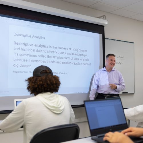 Assistant Professor of Management Thomas Lombardi stands in front of a projector screen facing a classroom of students with open laptops.