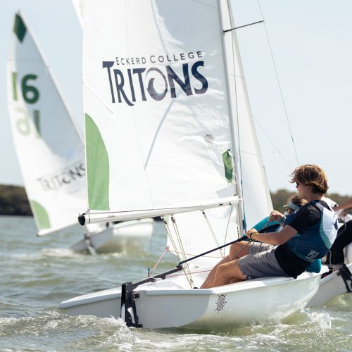 Two sailors on a sailboat with a sail that reads TRITONS