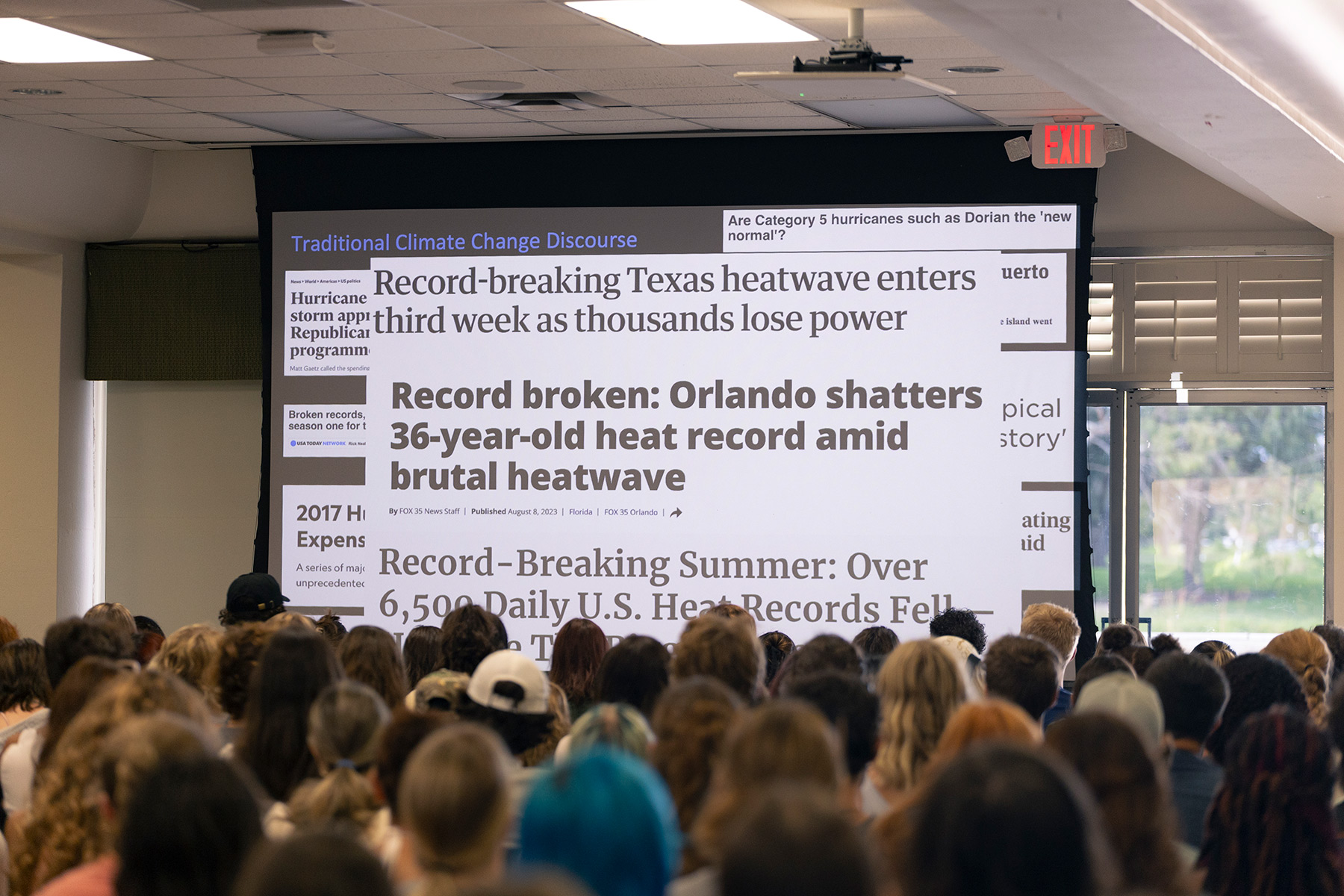 A large screen showing news clipping about broken heat records as a large audience looks on