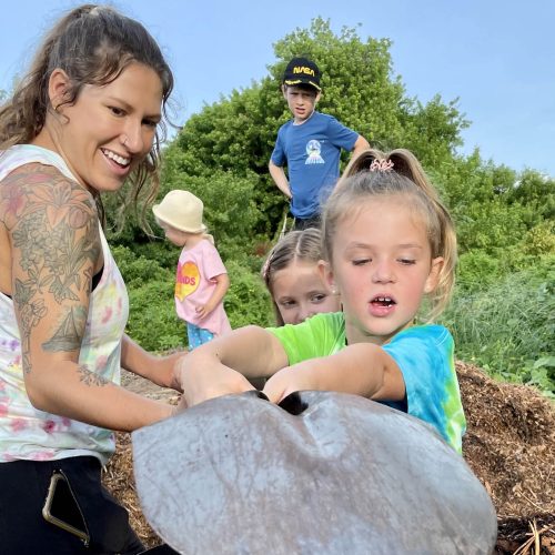 Young woman with tattoos helps a young girl use a shovel at a mulch pile