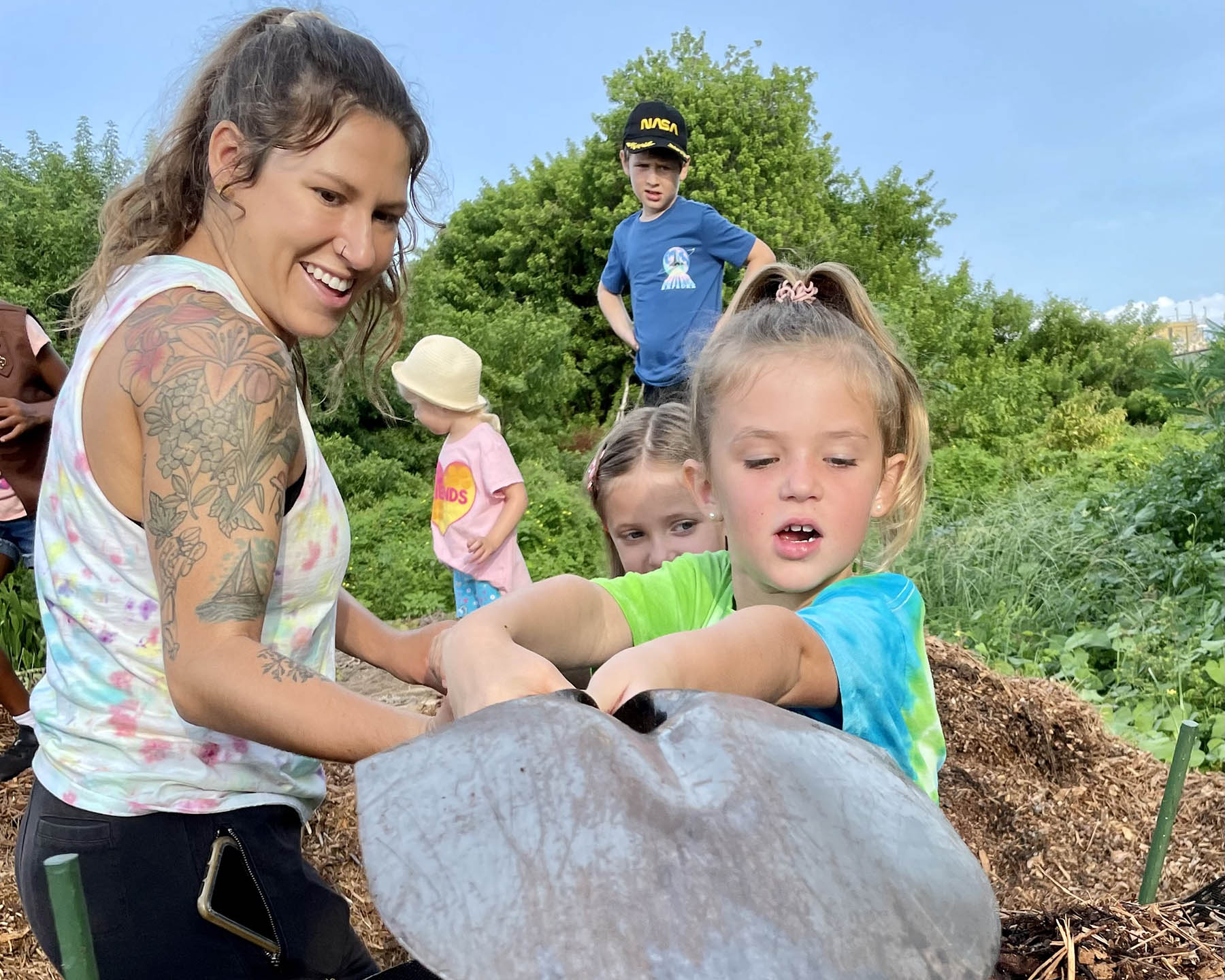 Young woman with tattoos helps a young girl use a shovel at a mulch pile