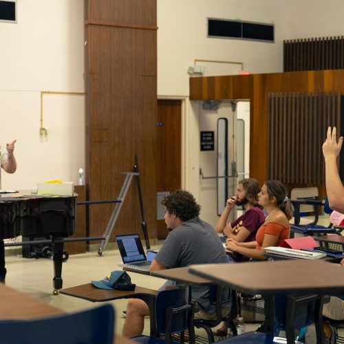 Professor stands behind a piano while pointing to students seated around him who are raising their hands