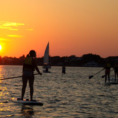 Paddleboarders and sailboat head out over the water towards an orange sunset