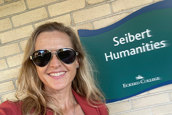 Woman in sunglasses in front of a brick wall with a sign that reads Seibert Humanities