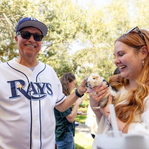 Man in Rays shirt and hat stands next to student holding guinea pig