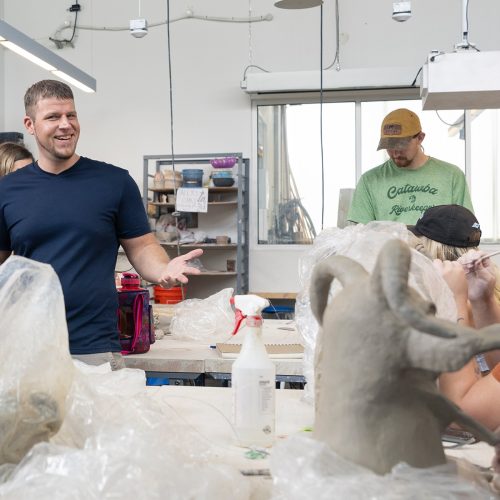 Professor smiles at a couple students seated at a big table with clay and clay tools