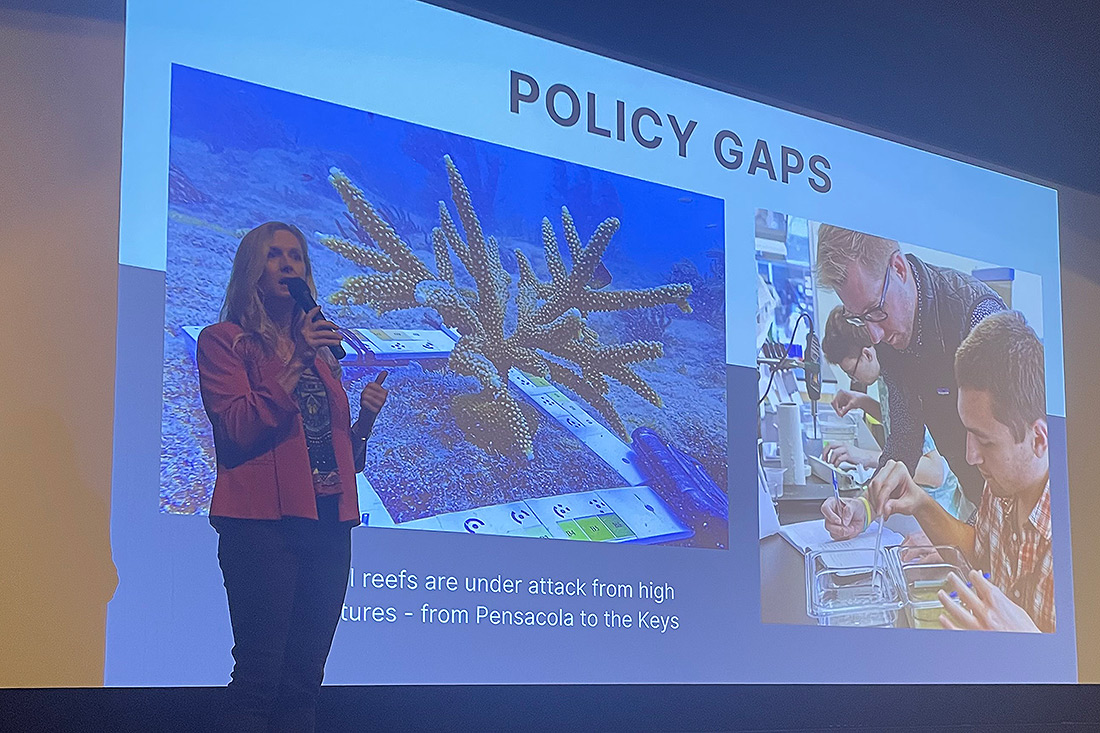 Woman on stage with handheld microphone and big screen behind her with a slide that reads "policy gaps"