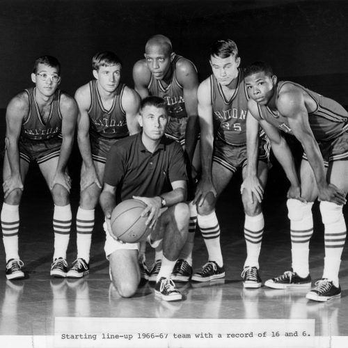 Five students with hands on knees in basketball attire stand around a coach holding basketball