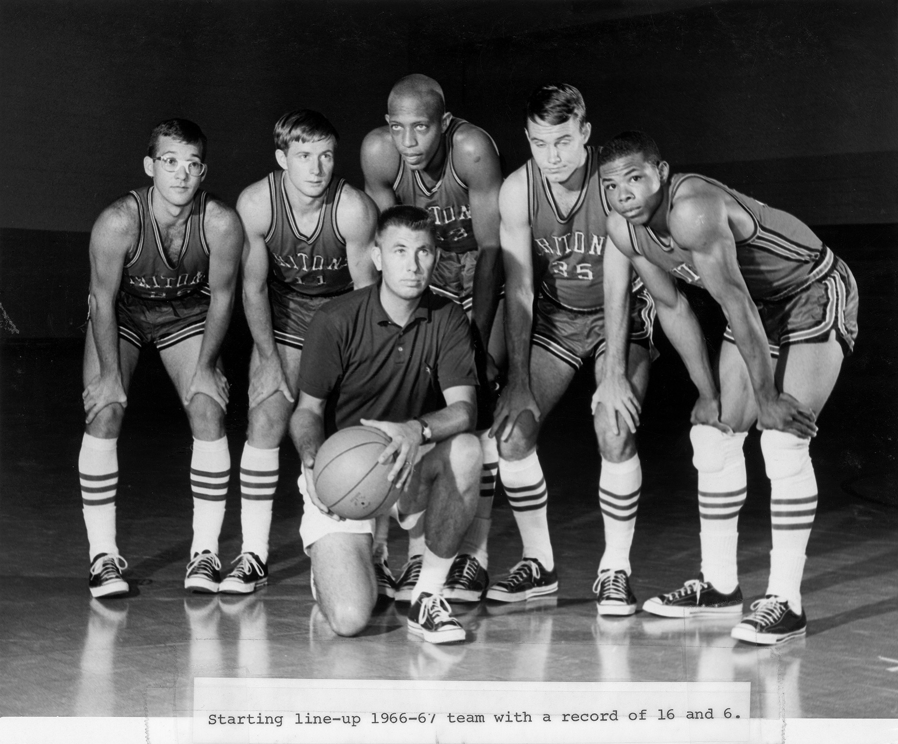 Five students with hands on knees in basketball attire stand around a coach holding basketball