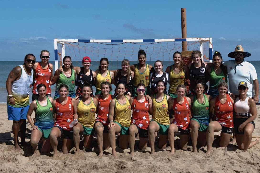 Group of about 20 female athletes and coaches posing on the beach in athletic gear with a large net behind them