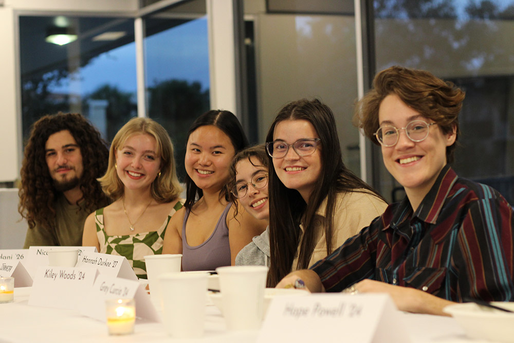 Six students smiling while seated side by side at a table with name placards