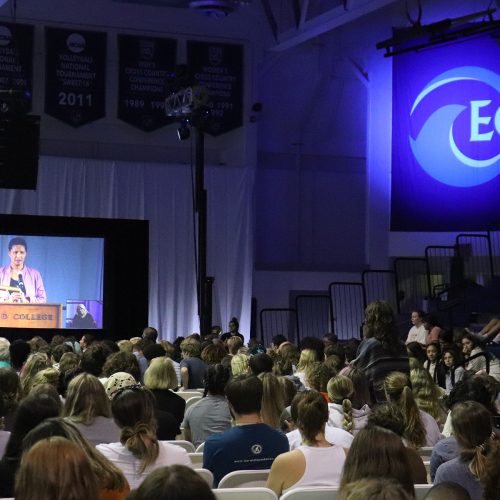 A speaker's image appears on a very large screen in a gym auditorium with hundreds of attendees seated and listening and a large banner that has the letters "EC"