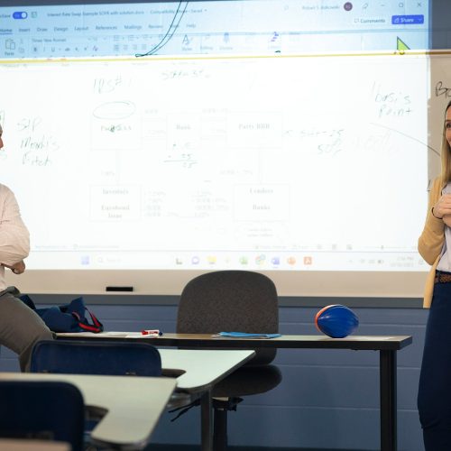 Professor sitting on edge of desk smiles at woman addressing the class