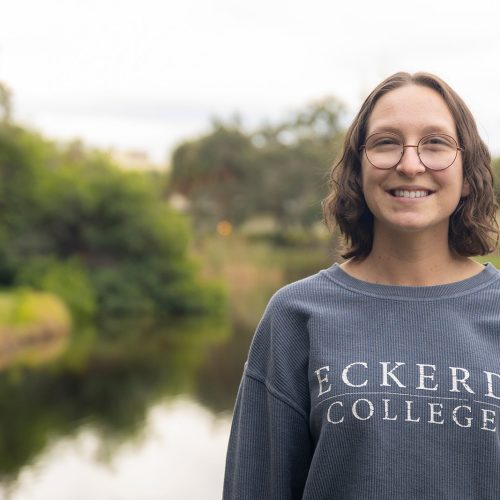 Student in glasses wearing a sweatshirt that says "Eckerd College" stands before a pond with mangroves near the shoreline