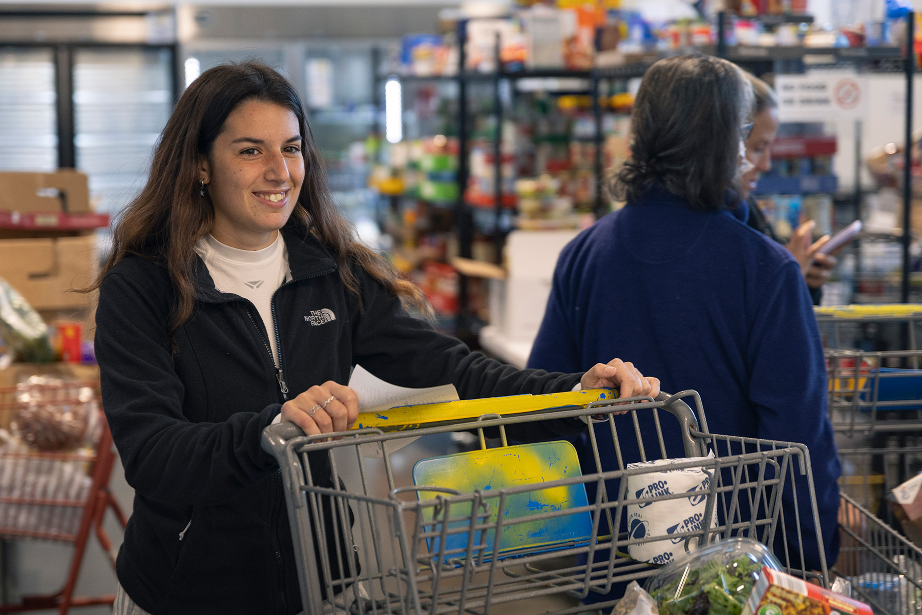 Student smiles while pushing a shopping cart of food inside a pantry