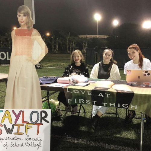 Cardboard cut-out of Taylor next to a table where three students sit with a sign "Love at first thigh"