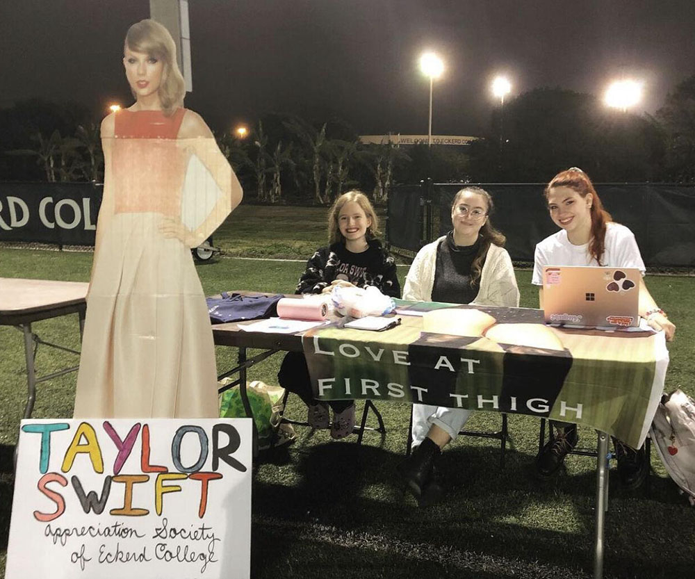 Cardboard cut-out of Taylor next to a table where three students sit with a sign "Love at first thigh"