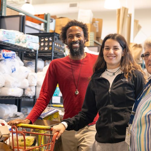 Two adults stand with an Eckerd student in a food pantry while pushing a shopping cart with food inside it