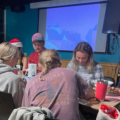 Students, some wearing Santa hats, sit around a table working on a group activity while a holiday movie plays o a screen behind them