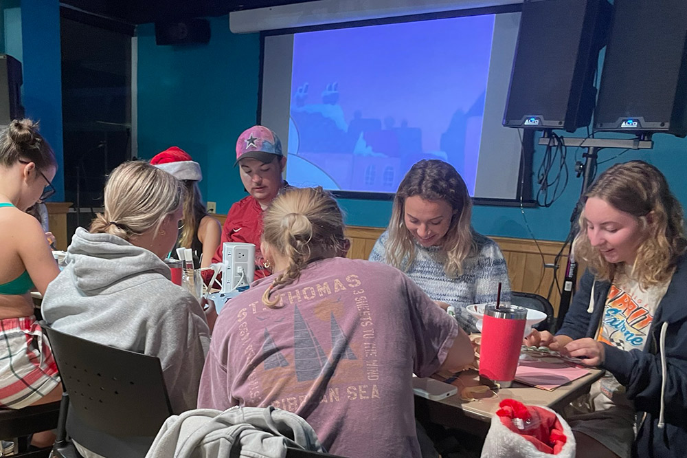 Students, some wearing Santa hats, sit around a table working on a group activity while a holiday movie plays o a screen behind them