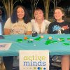 Three students smiling and seated at a table covered in blue cloth with a sign that reads "active minds"
