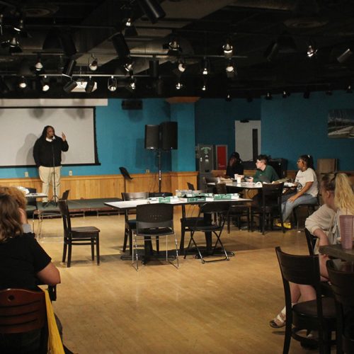 Room with speaker on stage and half a dozen tables with seated students