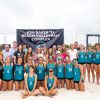 A large group of people including the beach volleyball team stands in front of a sign that reads Jon Baker '91 Beach Volleyball Complex