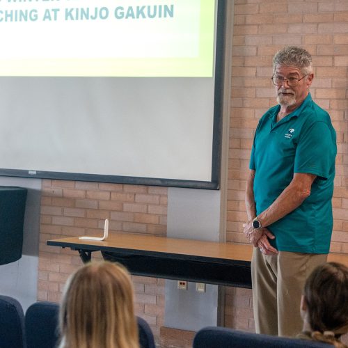 Man in teal shirt speaks before an audience of students