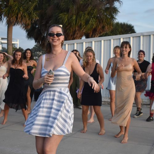 Student in sunglasses smiles at camera as group of students dances behind her