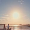 Two girls walking along bridge by large body of water while sun sets