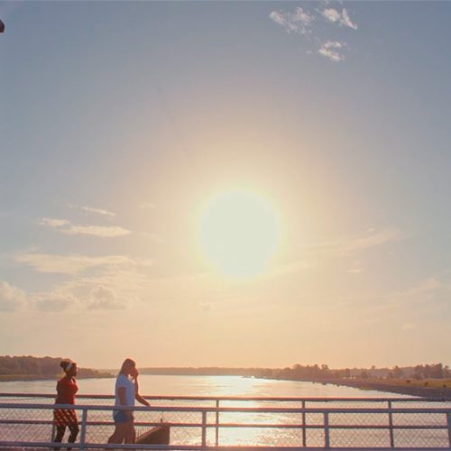 Two girls walking along bridge by large body of water while sun sets