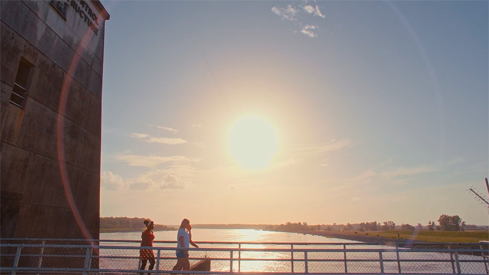 Two girls walking along bridge by large body of water while sun sets