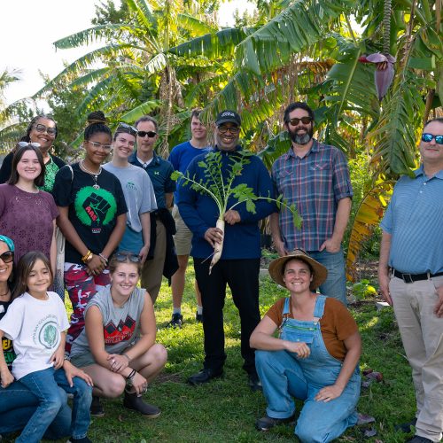 Group of students, staff and faculty stand with guest holding plant at a farm location