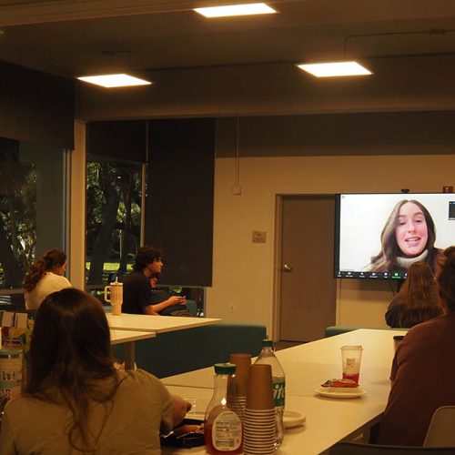 Students seated around tables to watch a big monitor at the front of the room showing a person speaking