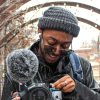 Student wearing hat adjusts his camera while smiling as the snow falls
