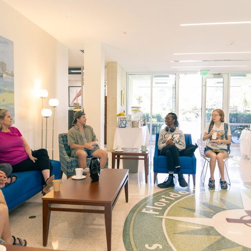 Students and staff sit on comfortable furniture to chat with the president, who stands and smiles