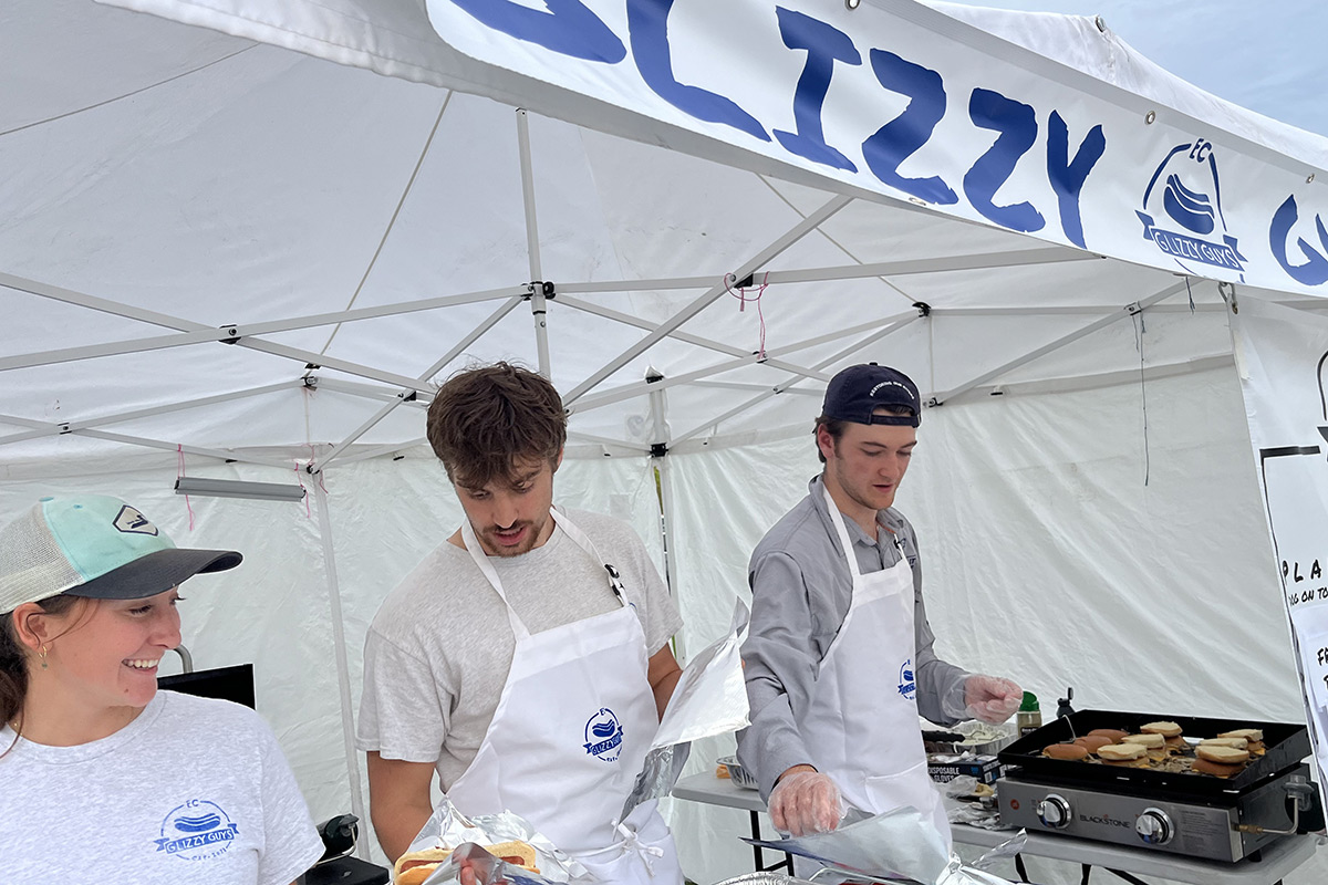 Three students wearing aprons serve hot dogs and burgers from under a tent that reads Glizzy Guys