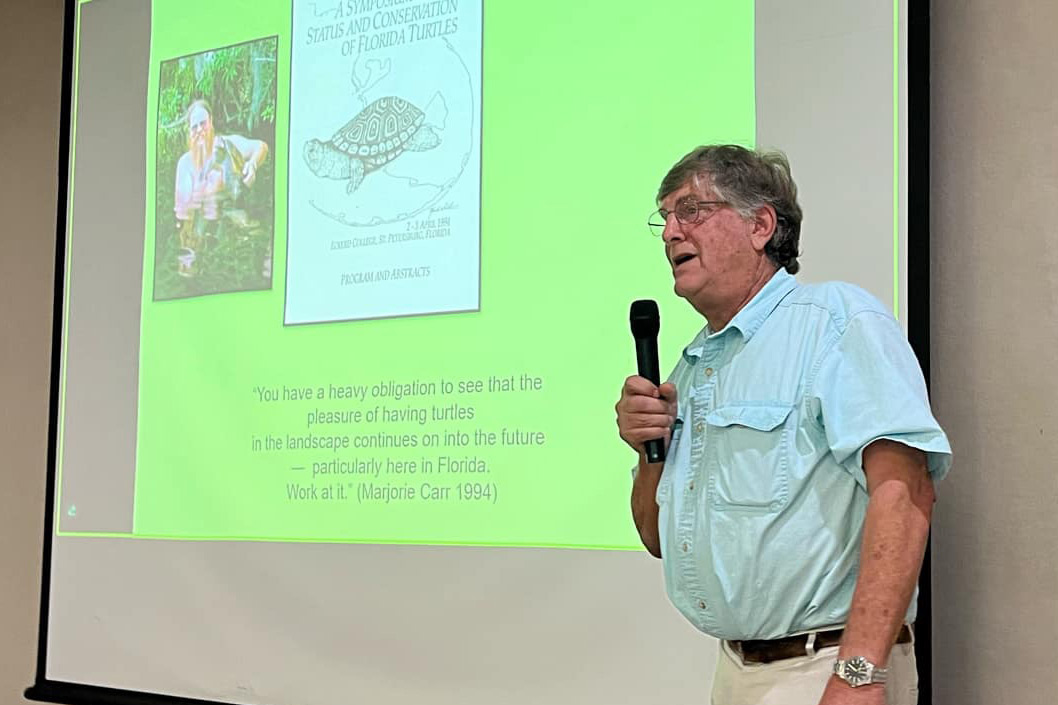 Man in glasses speaks into microphone in front of screen showing a photo of turtle researcher