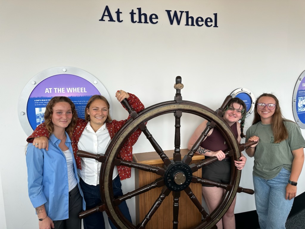 Four students stand by a large ship's steering wheel with "At the wheel" on the wall behind them