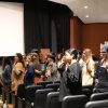 Group of students hold up one hand as they stand in the aisles of an auditorium as they listen to a speaker at a podium on stage