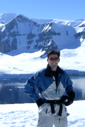 Student in sunglasses and winter gear stands in front of mountain range and icy water
