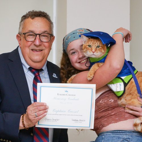 Student holds cat wearing graduation cap next to president who holds a certificate