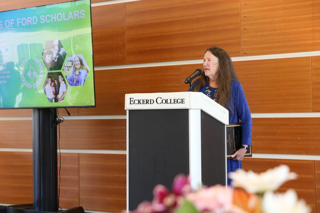 Woman at podium holding plaque and standing by monitor that reads FORD SCHOLARS