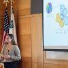 Student stands at podium in front of American flag and a large screen showing a slide of genetic illustrations